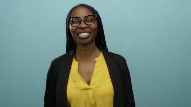 Sarı bir bluz içinde gülümseyen gözlüklü siyah kadın mavi bir duvarın arkasında duruyor..