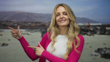 Sahilde pembe bir kazakla gülümseyen bir kadın manzaralı bir arka planı işaret ediyor..