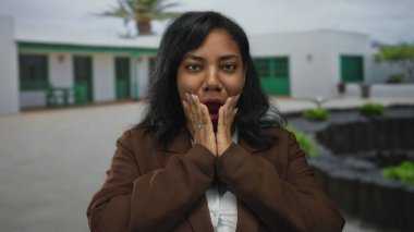 Elleri yanaklarında kahverengi ceket giyen bir kadın beyaz bir binanın önünde dikiliyor..