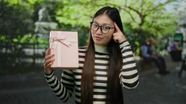Açık hava parkında pembe hediye kutusu tutan kadın uzun saçlı ve gözlüklü, yemyeşil ve halka açık bir alanda fıskiyeler arasında neşeli bir etki yaratıyor.