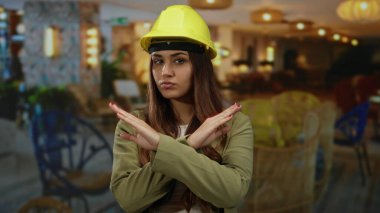 Sarı kasketli kadın mimar, şık otel odasında X hareketi yapıyor ve güçlü profesyonel duruşuyla reddediyor.
