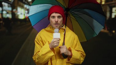 Gece sokağında renkli şemsiye altında mikrofon tutan genç muhabir sarı yağmurluk ve kırmızı bere giyiyor..