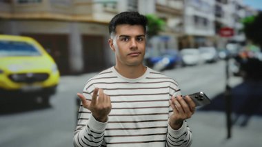 Şehir caddesinde akıllı telefonlu genç bir adam açık havada kargaşa yaratıyor arka planda bulanık araçlar ve binalar olan sıradan çizgili bir gömlek giyiyor..