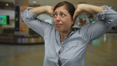 Orta yaşlı mavi çizgili gömlekli kadın havaalanı terminalinde kulaklarını gergin bir ifadeyle kapatıyor..