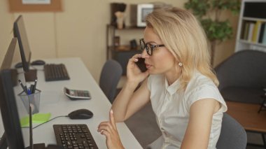 Ofis binasındaki beyaz bir masada klavye ve fare yazarken gözlük takan kadın telefonu kulağına tutuyor; konsantrasyon verimliliği.