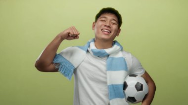 Futbol topu ve eşarpla gülümseyen genç adam izole edilmiş sarı arka plana karşı, spor için enerji ve coşku gösteriyor..