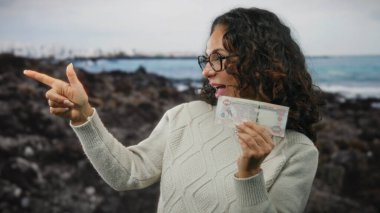 Kadın elinde su ve taşlarla kumsalda elindeki Dirham banknotuyla neşe dolu bir deniz kenarı anını göstererek mutlu mesut işaret ediyor..