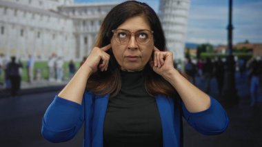 Kadın, gözlüklü ve mavi ceketli sidik kulesi binasının önünde parmaklarıyla kulaklarını kapatıyor; sinir bozucu seyahat hüsranı.