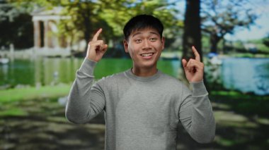 Genç adam gülümsüyor ve işaret ediyor, canlı yeşillik ve ağaçlarla dolu verimli bir açık hava parkında duruyor, mutluluk ve pozitifliği iletiyor..