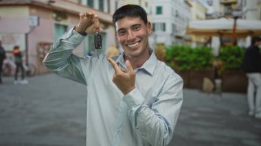 Genç gülümseyen adam arabanın anahtarını tutarken, onu güneş ışığında kaldırımda işaret ediyor; yeni sahiplik sevinci.