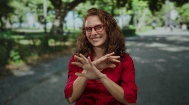 Güneşli bir parkta kırmızı gömlek ve gözlük takmış yeşil ağaçlarla çevrili açık havada eğlenen bir kadın.