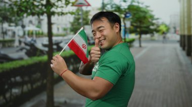Başparmaklarıyla gülümseyen genç adam İran bayrağını şehir caddesinde tutuyor.