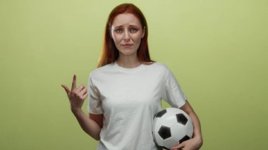 Sarı bir duvara karşı şaşkın bir ifadeyle futbol topu tutan bir kadın, izole bir arkaplanda kırmızı saç ve günlük beyaz gömlek.