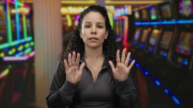 Genç İspanyol kadın, arka planında renkli kumar makineleri olan canlı bir kumarhanede sakin bir şekilde el kol hareketi yaparak heyecanın ortasında sükunet duygusunu iletiyor..