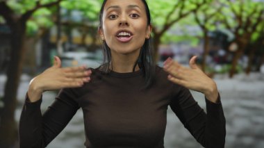 Canlı bir şehir caddesinde enerjik bir şekilde iletişim kuran bir kadın, enerjik ifadeler sergiliyor ve yeşil bir zemin arasında el hareketleri yapıyor..