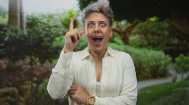 Kısa saçlı, beyaz tişörtlü bir kadın yemyeşil bir ormanda parmağını kaldırıyor, kendine güveni tam..