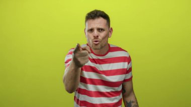 Parlak sarı arka planda, kolunda dövmeleri olan, çizgili tişört giyen, kararlılık odaklı ve nişanlı olduğunu gösteren bir adam.