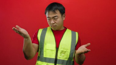 Yansıtıcı yelek giyen genç adam, kafa karışıklığı ve düşünceleri taşıyan canlı kırmızı bir zemin üzerinde duruyor..