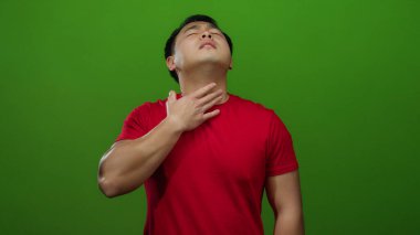 Kırmızı gömlekli, yeşil arka planda boğazı ağrıyan genç adam rahatsız ve ağrılı görünüyor, boynunu sıkıyor sanki rahatsızlık ya da acı çekiyormuş gibi..
