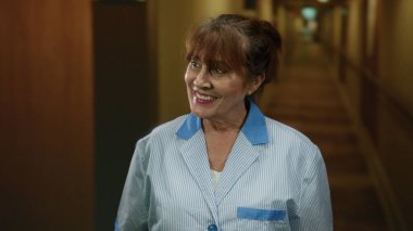 Gülümseyen kıdemli kadın otel koridorunda duruyor, ev ortamında konukseverliği ve profesyonelliği somutlaştırıyor..