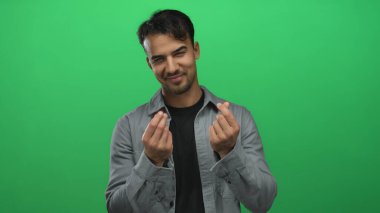 Genç İspanyol asıllı bir adam, yeşil bir arka plana parmaklarıyla para hareketi yapıyor. Günlük kıyafetleriyle neşeli ve kendinden emin bir tavır sergiliyor..