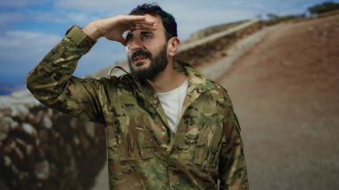 İspanyol üniformalı bir adam, dağlık doğal bir ortamda duruyor, gözlerini kapatıyor ve gülümsüyor, engebeli araziye kusursuz bir şekilde karışıyor..