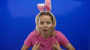 Pembe gömlekli ve tavşan kulaklı kadın, canlı mavi renkli stüdyoda parmak göğse işaret ediyor. Sürpriz!.