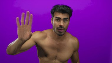 Göğüs kasları kaslı, Latin kökenli genç adam mor stüdyoda parmaklarını aşağıya doğru gösteriyor..