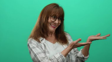 Kıdemli İspanyol kadın canlı yeşil bir duvara başparmağıyla gülümsüyor, gözlük takıyor ve günlük giysiler giyiyor, pozitifliğini ve dostluğunu iletiyor..