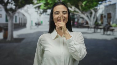 Güneşli bir sokakta, beyaz tişörtlü, sessiz bir kadın. Ağaçlar, sakin ve sakin bir hava sergiliyor..
