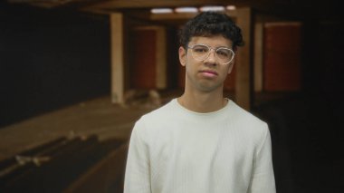 Gözlüklü adam kafası loş tamamlanmamış bina içinde eğik; belirsizlik içsel gerginlik şüphesi.