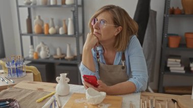 Kadın akıllı telefonu tutuyor ve çenesini ahşap bir bölmede seramik vazolarla çevrili çömlek atölyesinde kil kasesiyle çalışıyor..