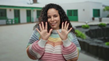 Arka planda evler, mutluluk ve doğal güzellik saçan günlük bir sokak sahnesinde renkli bir süveter giyen kadın..