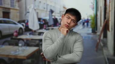 Genç bir adam açık hava restoranının terasında dikilirken düşünceli bir ifadeyle bulanık bir şehir geçmişine karşı.