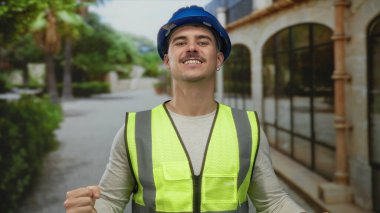 Yansıtıcı yelek ve kasketli genç İspanyol bir adam, şehir inşaat temalarını somutlaştıran bir caddede güven içinde duruyor..