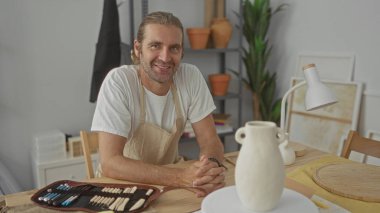Önlük takan sarışın adam, güneş ışığı altındaki bir stüdyoda, ahşap bir masanın üzerinde seramik bir vazonun yanında dinleniyor..
