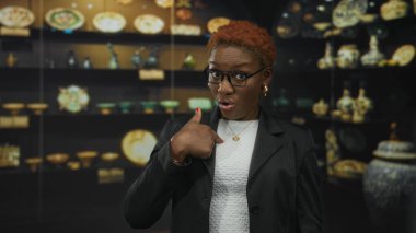 Gözlüklü, beyaz bluz ve kalp kolyesi takan kadın vazoyu işaret eden el kaldırıyor; şüphecilik.