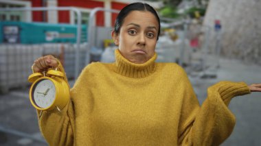 Sarı süveter giyen genç bir kadın. Elinde sarı alarm saati ve şantiyede omuz silken bir ifade var.