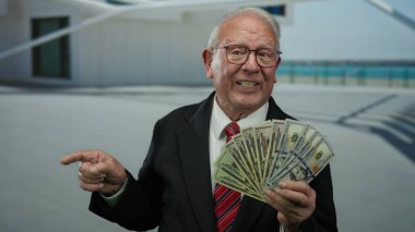 Kıdemli adam bize sokakta dolar tutuyor, iş kıyafetleriyle gülümsüyor, gün ışığı altında açık havada zenginlik ve başarı sergiliyor..