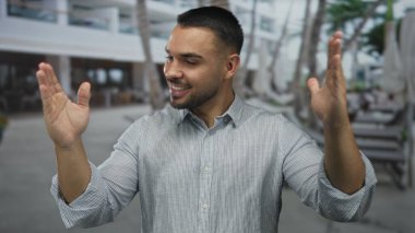 Çizgili gömlekli bir adam kollarını modern beyaz bir binanın yanında palmiye ağaçları ile geniş bir caddeye yayar; sıcak karşılama neşe.