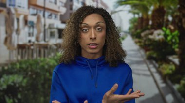 Meşgul caddede palmiye ağaçları ve yeşil çalılarla kaplı mavi bir kapüşonlu kadın jestleri; kendine güven.