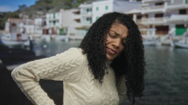 Beyaz kablo örgü süveterli kadın, mavi suyun yanında kıyıya yanaşmış tekneleri olan bir bina tarafından sırtından tutuluyor..