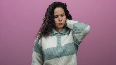 Üzüntülü bir ifadeyle pembe arka planda duran bir kadın, sanki rahatsız olmuş gibi boynunu tutuyor, çizgili bir kazak giyiyor, stresi veya gerginliği vurguluyor.