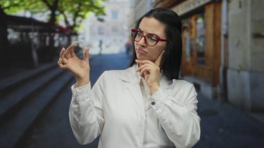 Genç bir dişçi, görünmeyen diş tellerini düşünceli bir şekilde bir sokakta tutuyor, üniforma giyiyor, şehirde odaklanmış bir ifadeyle ortodontik bakım sergiliyor..