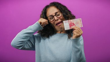 Orta yaşlı bir kadın elinde İzlanda Krona banknotuyla canlı mor bir arkaplan tutarken komik bir şekilde ağlıyor. Finansal kaygılarını belli ediyor ya da sahte bir sıkıntı yaşıyor..