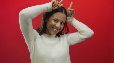 İspanyol kadın gülümsüyor ve parmaklarıyla neşeli bir jest yapıyor cesur kırmızı arka plan duvarına karşı neşeli ve enerjik bir duruşta boynuzları andırıyor..