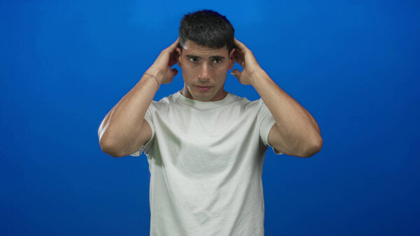 Молодой латиноамериканец приспосабливает волосы руками возле ушей в синей студии, глядя вперед; концентрация.