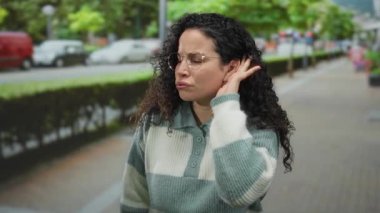 Kıvırcık saçlı ve gözlüklü İspanyol kadın sokakta dikiliyor, eli kulağında, yeşillik ve araçlarla çevrili bir şekilde dinlemek için işaret ediyor..