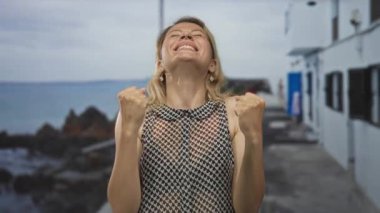 Kadın, kayalık kıyılara ve okyanus dalgalarına tepeden bakan beyaz bir sahil binasının yanında yumruklarını kaldırır. Zafer zaferi zaferi kutlar mutluluğu..