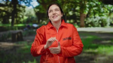 Turuncu hapisteki kadın dışarıda yeşil ve güneş ışığıyla çevrili bir parkta özgürlük ve hapsedilme arasında bir zıtlık hissi yayıyor..